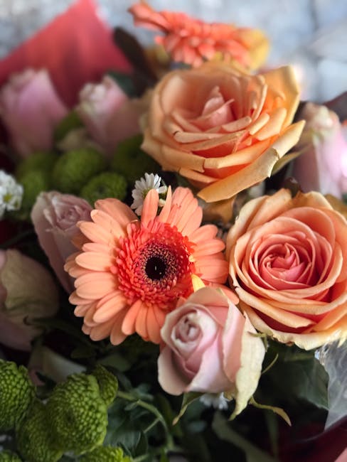 A close-up view of a vibrant floral arrangement featuring peach-colored roses with tightly layered petals and a soft sheen, complemented by a bright pink gerbera daisy with a dark center, alongside sm