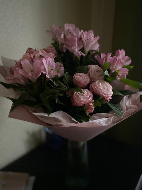 A bouquet of fresh pink flowers, including roses and lilies, arranged with dark green foliage and wrapped in light pink tissue paper. The bouquet is displayed on a dark surface in an indoor setting wi