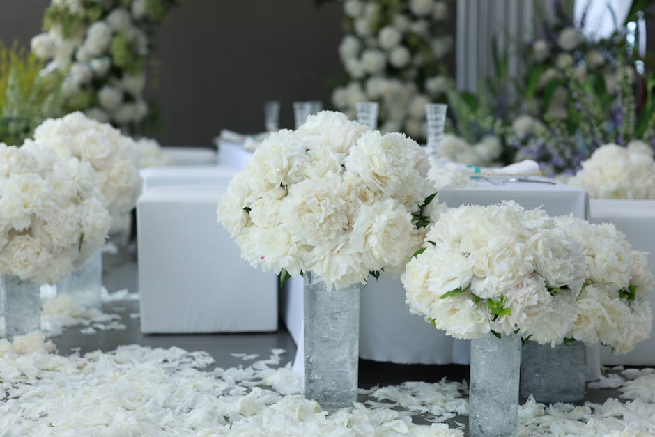 A floral arrangement featuring large, rounded bouquets of white hydrangeas and roses, placed in tall, rectangular, concrete vases and arranged on a table covered with scattered white flower petals. Th
