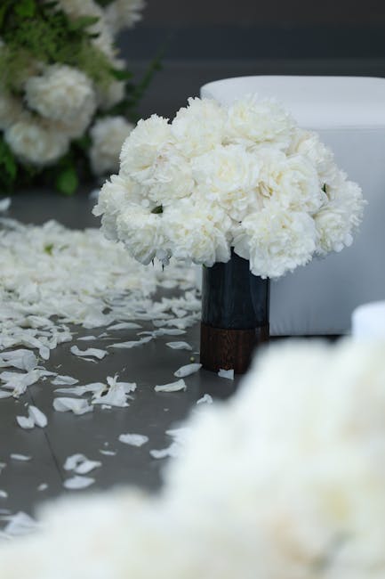 A round floral arrangement of fresh white carnations, densely packed to form a voluminous cluster, sits in a modern black and brown vase on a table covered with white petal confetti. The bouquet is pa